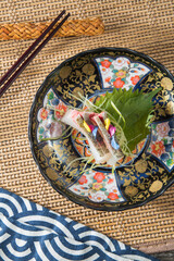 A decorative plate with gold and floral patterns presents delicate sashimi slices with shiso leaf and edible flower petals. Placed on woven mat with chopsticks for an elegant overhead composition.