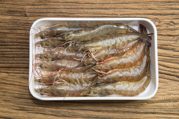 A white tray holds neat rows of raw shrimp with shells and antennae intact on a rustic wooden surface. Moist translucent bodies signal freshness, ready for cooking.