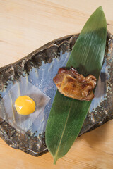 A small grilled meat piece is plated on a leaf over a ceramic dish with a glossy raw egg yolk on the side. Minimalist styling and warm lighting evoke refined Japanese dining.