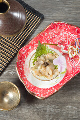 An ornate red dish holds a small bowl of marinated seafood topped with roe, shiso leaf and a flower. Lacquer ware and woven mats complete the elegant overhead setting.