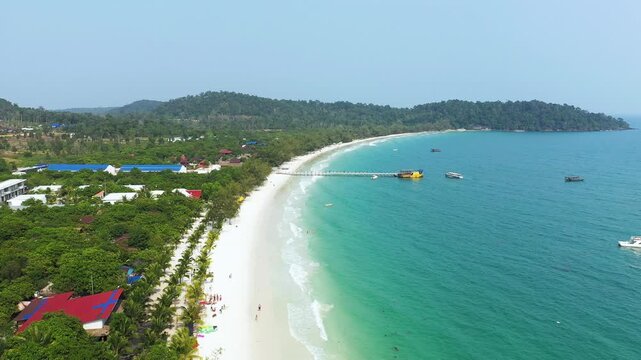 Scenic aerial view of Long Set Beach with turquoise water, white sand, and lush greenery. Boats are anchored near a wooden pier extending into the sea.
