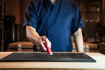 A chef places a piece of tuna nigiri on a dark board at the counter. Soft light emphasizes the red fish and tidy rice, capturing an artisan service moment in a traditional setting.
