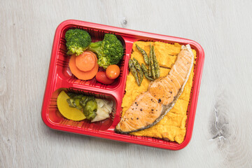 A red bento tray holds a grilled salmon fillet, layered omelette, broccoli, carrot, cherry tomato and pickles. The clean overhead view suggests a convenient, balanced Japanese style meal.