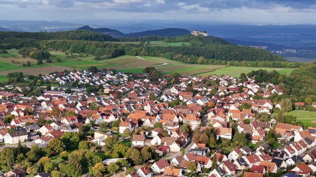 Aerial view of Neuffen, showing the vibrant red-tiled roofs of homes contrasting with green trees and the distant Hohenneuffen Castle, Erkenbrechtsweiler, Baden-Wurttemberg, Germany.