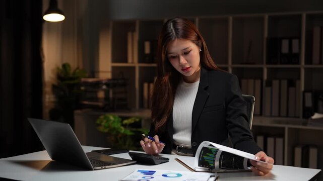 Young Asian accountant calculating data from financial documents and charts while working overtime at her desk in a dark office, committed to meeting a critical company deadline
