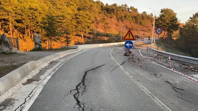 Severe Road Collapse. A fractured asphalt road with a deep drop, marked by hazard tape and a blue arrow sign, set against a sunlit hillside. 