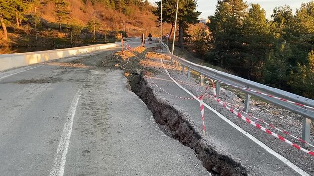 Severe Road Collapse. A fractured asphalt road with a deep drop, marked by hazard tape and a blue arrow sign, set against a sunlit hillside. 