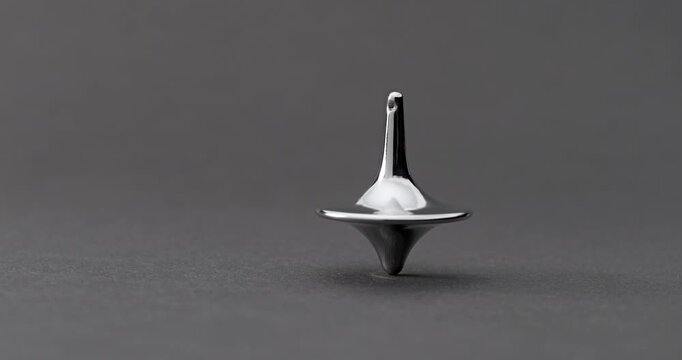 Metallic Spinning Top Falling Over. Linear footage of a shiny chrome top in steady rotation, progressively wobbling with increasing tilt until it fully topples on a matte gray surface.