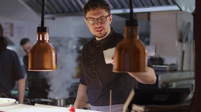 Professional chef placing finished plate on counter, ringing bell, then reading order ticket and assigning task to coworkers, coordinating food preparation workflow in restaurant kitchen