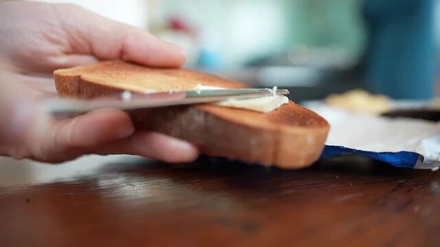 Hand holds toasted bread while adding a pat of butter with a knife over a wooden table, simple homemade breakfast preparation. Close-up food b-roll.