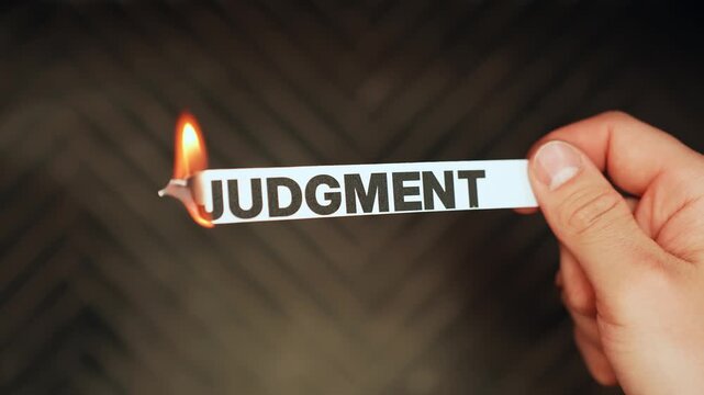 Close-up of a hand holding a paper strip labeled &ldquo;JUDGMENT&rdquo; as the edge burns with flame on a dark background. Letting go of criticism and shame concept with copy space.