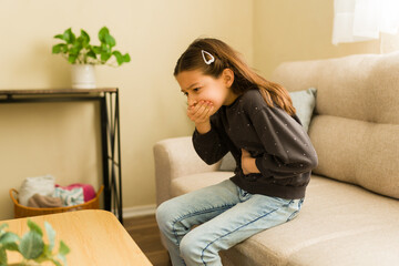 Little girl sitting on a sofa, clutching her stomach and covering her mouth, feeling unwell with...