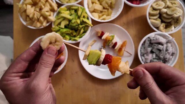 Men's hands string pieces of fresh fruit on a wooden skewer to make fruit kebabs, close-up