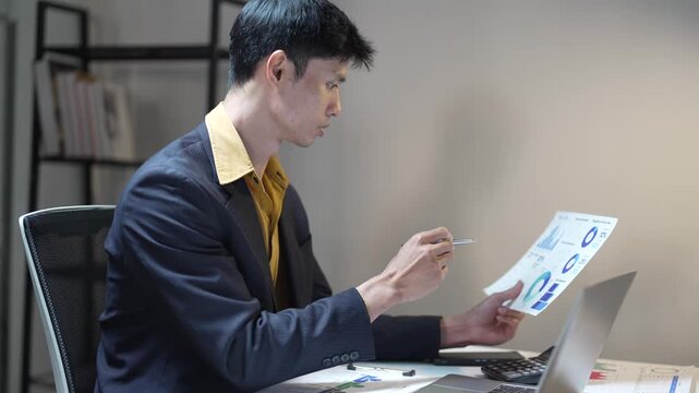 Focused asian businessman analyzing financial charts and graphs on documents and a laptop, working overtime in a modern office to meet deadlines and review project statistics for growth