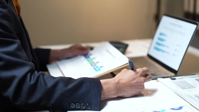 Diligent businessman sitting at a desk analyzing company financial reports, checking charts on documents and a laptop, and taking notes on a clipboard to develop a marketing strategy