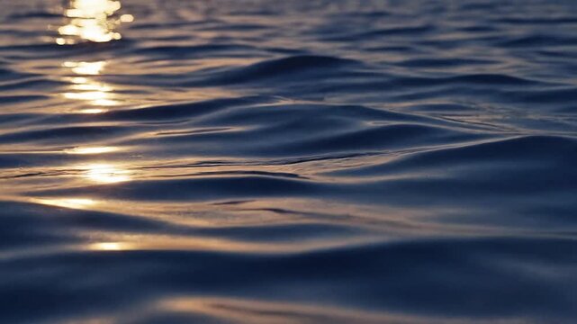 Tranquil dark blue water surface with gentle ripples reflecting the golden glow of the setting sun, creating a peaceful and serene natural abstract texture