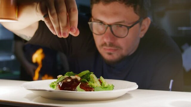 Professional chef sprinkling pine nuts over plated duck leg and vegetables, finishing dish under heat lamp in restaurant kitchen