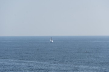 Fototapeta premium Sailboat gliding across the calm ocean waters under a clear blue sky. Yacht with white sails vast expanse of blue water. Background with faint outline of distant land, creating a serene maritime scene