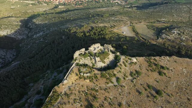 Drone shot of the ancient stone ruins of Chateau de Opoul Perillos surrounded by rugged landscape and distant village in southern France.