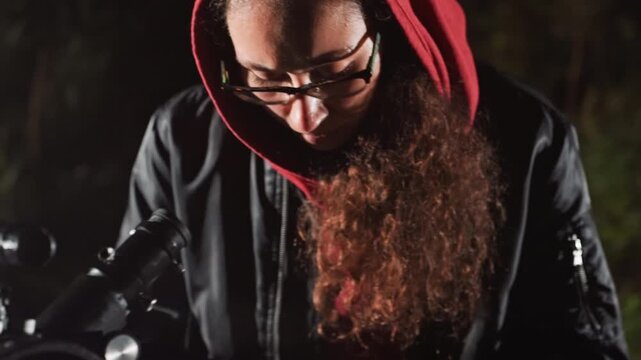Night backyard telescope observation observer in red hoodie leans over eyepiece, adjusting focus and scanning star field. Glasses reflect faint sky while hands finetune mount and camera rig. Mood