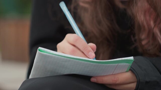 student writing notes with blue pen on spiral notebook outdoors, long hair, cozy coat, pearl bracelet, closeup hands making thoughtful gestures, focused study vibe under soft natural light