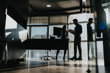 Fototapeta premium Two business professionals review documents in a modern office as sunlight fills the space. Colleagues stand near a desk with a computer, discussing work.