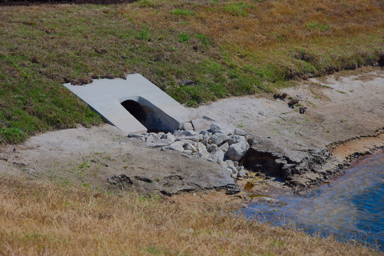 Concrete Culvert Drain at Rocky Shoreline Retention Pond  With Erosion and Runoff Near Dry Grassy Bank rock riprap on a dry grassy bank beside a shallow shoreline, showing erosion, stormwater runoff a