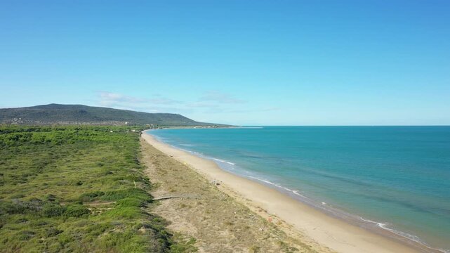 Aerial view of the pristine sandy beach and turquoise sea along the Isola Varano nature reserve in Italy, bordered by lush green vegetation under a clear blue sky.