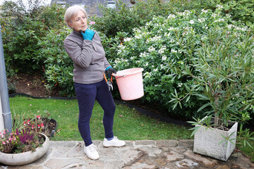 A retired gardener woman , thinks about her autumn tasks, stands in a magnificent garden, pruning shears and a bucket in her hand. She is surrounded by lush vegetation and colorful flowers. © Aygul Bulté