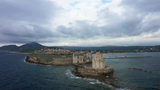 Dramatic aerial view of Methoni Castle on the rocky coastline of Methoni, Greece, with the sea and village in the background under a cloudy sky.
