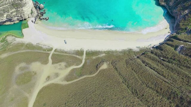 Aerial view of turquoise ocean waves meeting white sand between steep rocky cliffs and dirt paths at Afoul Beach, Salalah, Oman.