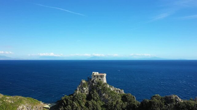 Scenic view of a historic stone tower perched on a rocky cliff above the deep blue sea at Capo di Conca on the Amalfi Coast, Italy. Lush greenery and clear skies complete this tranquil Mediterranean