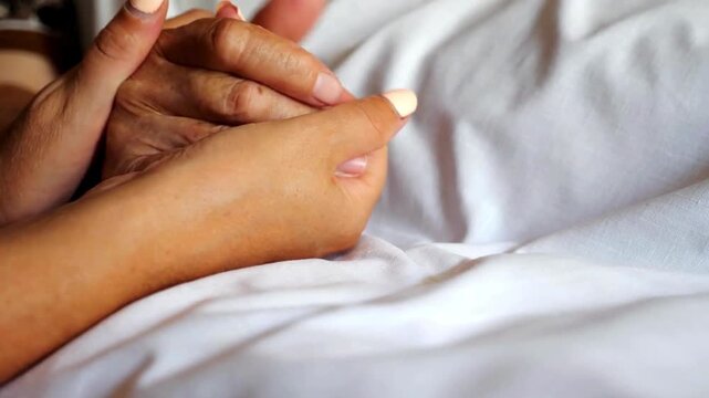 Unrecognizable worried woman gently stroking hand of her sick mother. Daughter comforting wrinkled arm of elderly mom that lying at bed in hospital.