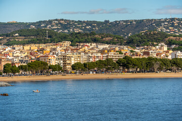 Fototapeta premium Sant feliu de guixols showing its vibrant beachfront promenade, sandy beach, and residential buildings climbing the green hills overlooking the calm mediterranean sea