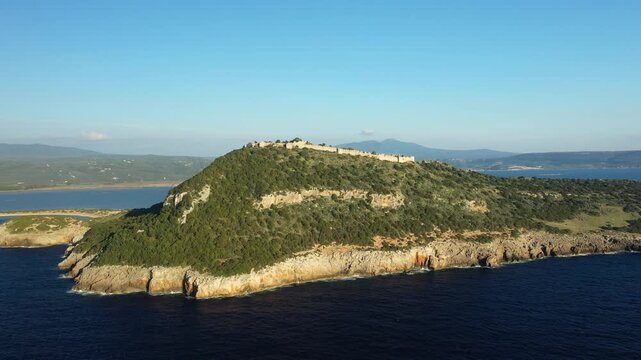Aerial view of Navarino Castle perched atop a lush green hill overlooking the sea in Greece. Rugged coastline and distant mountains complete the scenic landscape.