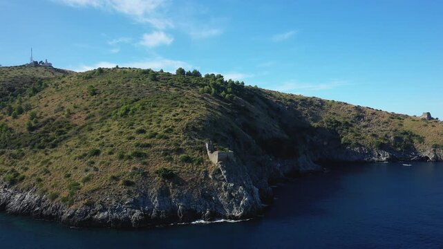 Aerial view of the rugged coastline and clear blue sea at Cap Palinuro, Italy, with lush green hills and rocky cliffs under a bright sky.