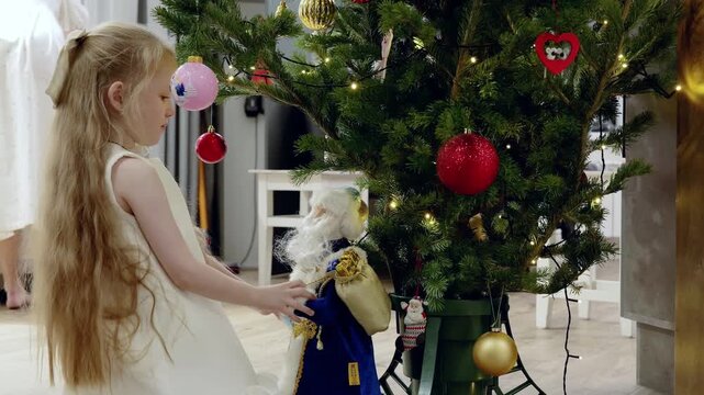 Blond girl in white dress gently adorning christmas tree, carefully placing nutcracker ornament with festive smile, creating warm holiday atmosphere at home