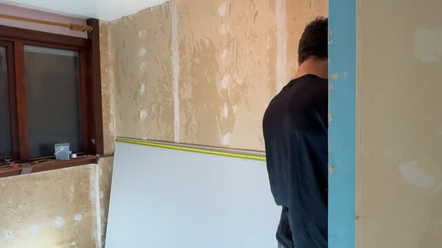 A man measures a sheet of drywall with a tape measure. Home renovation. Wall restoration. A builder's workflow. Repair work.