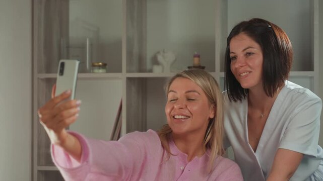 smiling client esthetician taking selfie after microcurrent facial in cozy treatment room, client in pink robe, esthetician in uniform, celebrating glowing skin, warm ambient lighting, shelf decor,