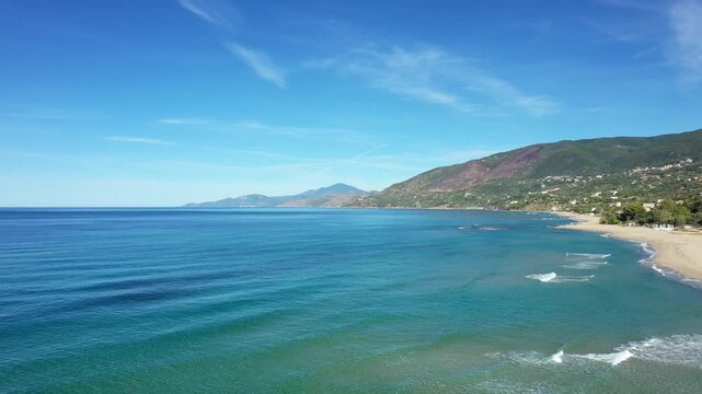 Scenic aerial view of the clear blue sea and sandy coastline near Palinuro, Italy, with lush green hills in the background under a bright blue sky.