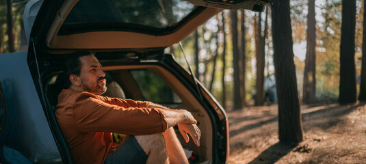 A young man is sitting relaxed alone in the open trunk of a car in the woods by the lake at sunset. © Anna