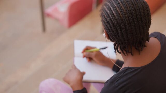 black student writing in notebook on bleachers, earbuds in, cornrows hairstyle, natural light, focused expression, pen movement, casual clothing, outdoor campus atmosphere, planning notes, solitude