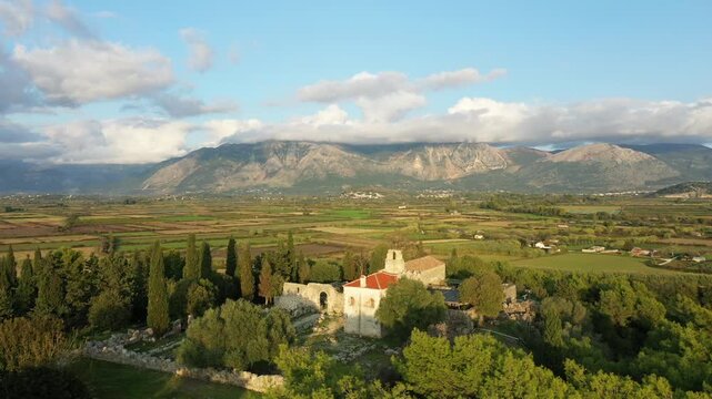 Aerial view of ancient stone ruins surrounded by lush greenery and dramatic mountains in Epirus, Greece, under a partly cloudy sky.