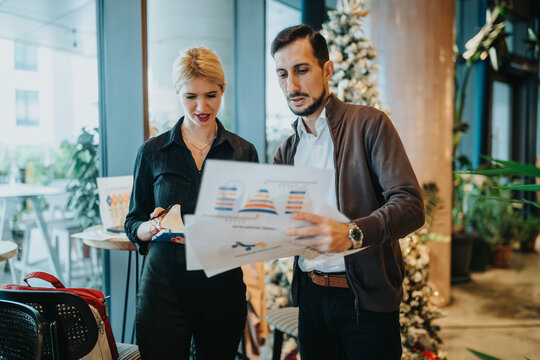 Two business partners study a charted report at a contemporary office. They discuss the data beside a Christmas tree with festive decor.