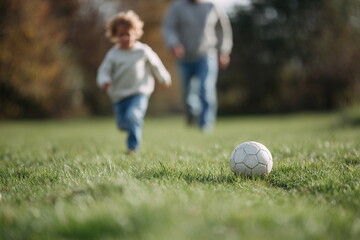 Obraz premium Young child running across grassy field toward a soccer ball with parent in background, capturing playful family moments outdoors