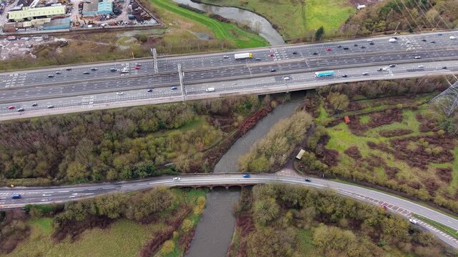 Aerial drone view of two bridges crossing a river near Birmingham, UK, contrasting a busy motorway with a small rural single-track road.