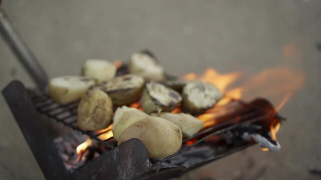 Potatoes are being grilled on a metal grate above a lively fire, capturing the essence of outdoor cooking. The flames and rustic setup evoke warmth and a sense of culinary adventure
