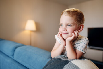 Fototapeta premium A heartwarming shot of a small boy leaning on a sofa with his chin on his hands, looking away with a sweet smile. Represents childhood curiosity, imagination, and peaceful home atmosphere.