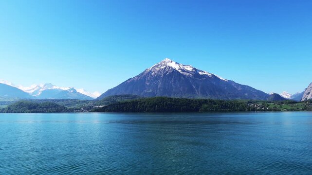 Cruising trip in Thun lake Switzerland, Snow mountain peak on clear blue sky with morning sunlight n sun light ray over calm water wave n forest reflection in spring season or springtime of Europe vdo