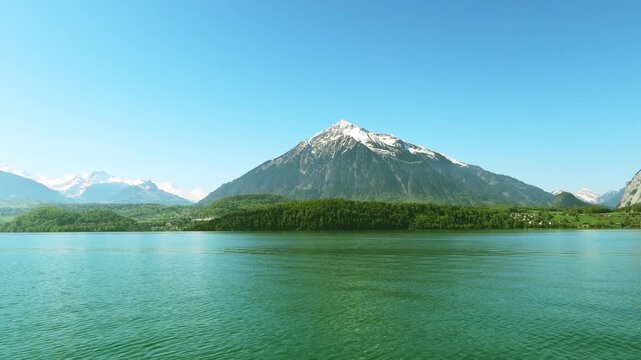 Spring shot of snowcapped or white snow mountain peak range in beautiful morning clear blue sky over calm water wave n green forest at Interlaken Lake Thun Switzerland, 4k aerial view, ferry boat ship